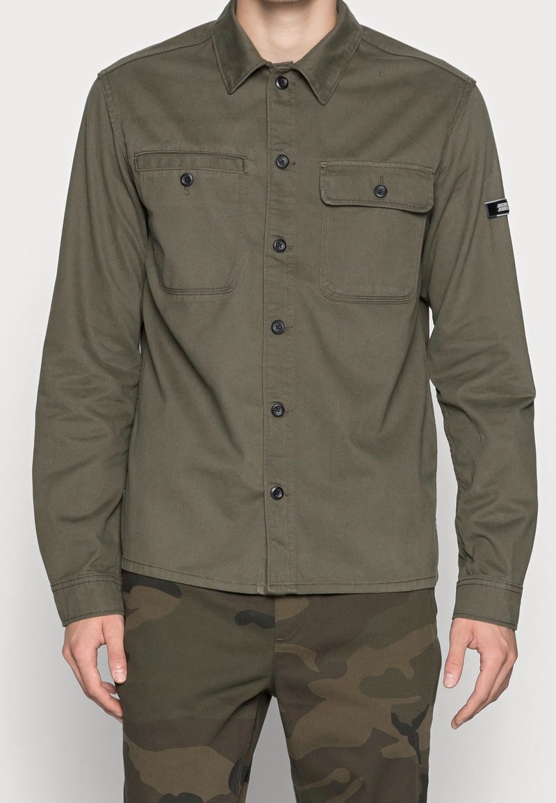 Uomo che indossa una camicia verde oliva con bottoni e due tasche sul petto, con pantaloni camouflage, in piedi con le braccia rilassate lungo i fianchi.