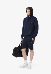 Felpa blu marina con tasca frontale, caratterizzata da un logo verde; abbinata a pantaloni corti blu marina e sneakers bianche. Borsa a tracolla nera tenuta in una mano.