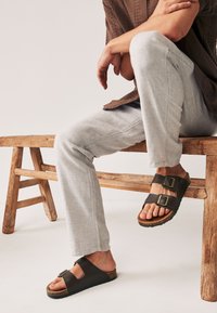 Sandales noires à deux brides avec boucles, semelle intérieure en liège et semelle en caoutchouc. Portées avec un pantalon gris clair, assis sur un banc en bois.