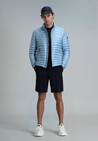 Doudoune bleu clair avec des coutures horizontales, fermeture éclair à l'avant et poches latérales. Portée sur une chemise noire et associée à un short bleu marine et des baskets blanches.