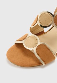 Tumman ruskea kiinni oleva sandaali, jossa on mokkanahkainen pinta, pyöreät kultaiset yksityiskohdat ja sileä sisävuori. Vankka pohja minimalistisella muotoilulla.