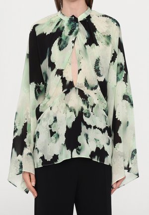 Woman wearing a loose, long-sleeve blouse with green, black, and white floral pattern and keyhole neckline paired with black pants.