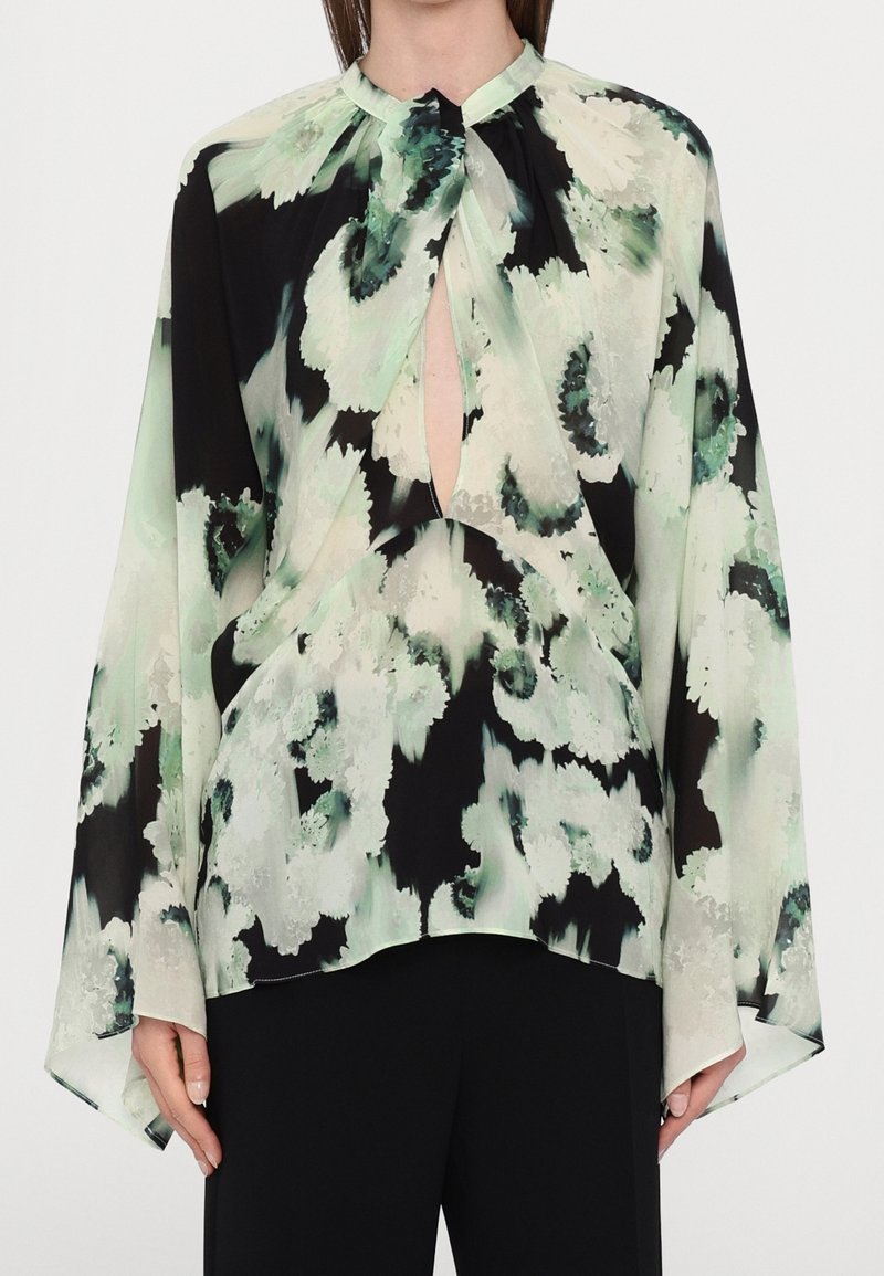 Femme portant un chemisier ample à manches longues avec un motif floral vert, noir et blanc et un décolleté en forme de clé, associé à un pantalon noir.