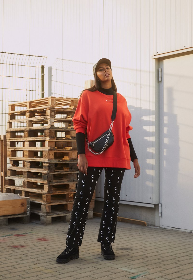Sweatshirt rouge surdimensionné, pantalon noir à motifs avec motifs d'yeux, sac bandoulière noir avec chaîne, et bottes noires épaisses. Palettes en bois en arrière-plan.