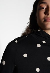 Zwarte stoffen top met witte stippen, met een hoge halslijn en een zachte textuur. Het ontwerp bevat schouderdetails zonder zichtbare sluitingen.