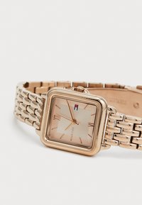 Montre carrée en ton or avec un cadran crème texturé, chiffres romains et un bracelet à maillons. Présente un logo minimaliste et une couronne.