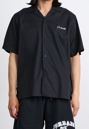 Person wearing a black short-sleeve button-up shirt with a small white "Carhartt" logo and black shorts with white "Jordan" text.