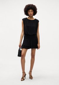 Vestido preto com decote franzido, mangas curtas e bainha em camadas e larga. Combinado com sapatos de salto fino e uma pequena bolsa com tachas.