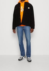 Svart manchesterjacka med framfickor över en orange hoodies, blå jeans och svart-vita sneakers. Avslappnad lager-på-lager-outfit.