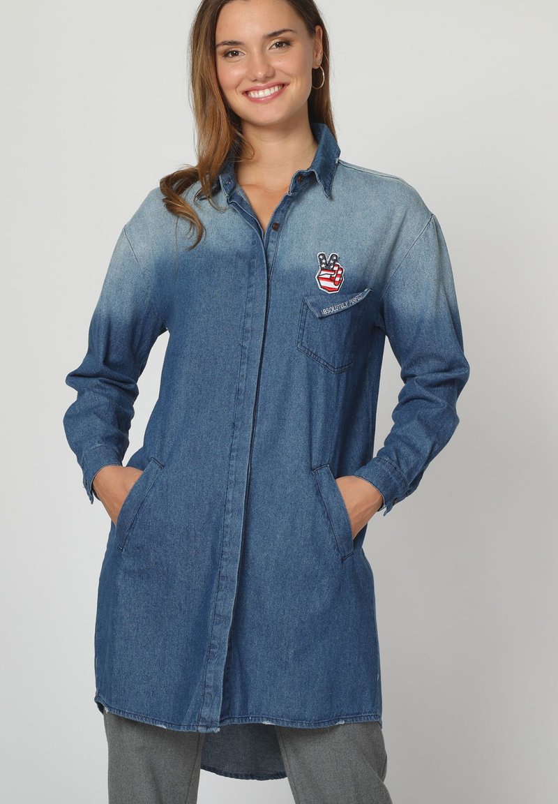 Mujer sonriente vestida con una camisa de mezclilla larga y en degradé, con bolsillos y un parche de signo de paz en el pecho, con las manos en los bolsillos, de pie contra un fondo gris.