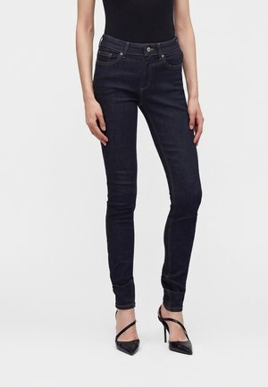 Femme portant un jean skinny bleu foncé et des chaussures à talons hauts pointues noires avec des brides croisées, debout devant un fond uni.