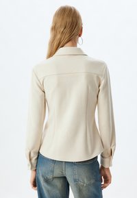 Camisa beige de manga larga con botones y cuello, que presenta un diseño ajustado y un ligero brillo, combinada con unos vaqueros azules de cintura alta.