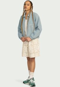 Pruhovaná modro-biela bunda, béžové krátke nohavice s vzorom, biela tielko, zelené tenisky, s dlhými zapletenými vlasmi a neutrálnym pozadím.