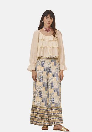 Blusa cremosa de manga larga con volantes y botones, combinada con pantalones anchos amarillos y azules con patrón y acentos en el borde. Sandalias casuales.