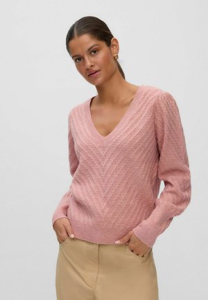 Femme aux cheveux foncés en queue de cheval basse, portant des boucles d'oreilles anneaux dorées, un pull rose texturé à col en V et un pantalon beige avec les mains dans les poches.
