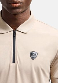 Polo beige con colletto zip; presenta un logo EA7 nero e bianco sul lato sinistro del petto; texture liscia, maniche corte.