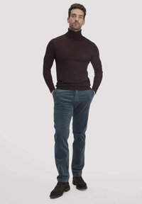 Maroonfarbener Rollkragenpullover kombiniert mit hellblauen Cordhosen. Akzentuiert mit dunklen Schuhen, vor einem neutralen Hintergrund stehend.