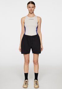 Canottiera beige senza maniche con accenti laterali blu; pantaloni corti neri con cintura elastica; calzini neri alla caviglia; scarpe beige multicolori.