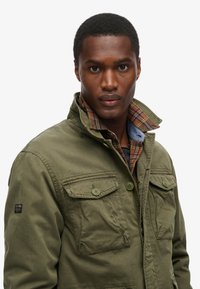 Feltjakke i olivengrøn bomuld med to brystlommer, knaplukning og en plaidforet krave i rige jordfarver.