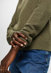 Pull en maille texturée vert olive avec des poignets côtelés, porté sur un denim bleu, mains avec une bague en argent retenant doucement la manche.