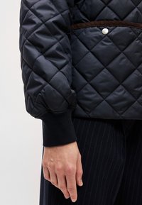 Melna džaka ar quilted (izšūtiem) rakstiem, kas iekļauj brūnu jostas detaļu un sudraba pogu, ar ribotām melnām aprocēm un gludu apdari.