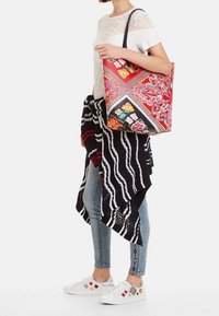 Sac fourre-tout rouge à motifs floraux et géométriques, avec des poignées noires et une forme rectangulaire structurée porté par une personne.