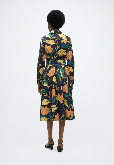 Femme debout de dos, portant une robe midi noire à fleurs jaunes et orange, associée à des sandales à talons noires.