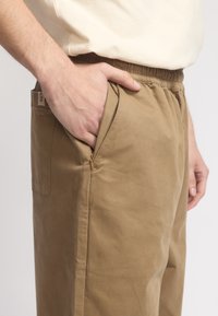 Katoenen tan broek met een relaxte pasvorm, elastische tailleband, zijzakken en subtiele stiksels. Het materiaal heeft een soepele textuur.