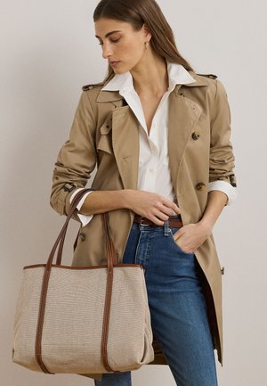 Kvinna som bär en beige trenchcoat och vit skjorta, håller en stor beige tygväska med bruna läderremmar, stående mot en enfärgad bakgrund.