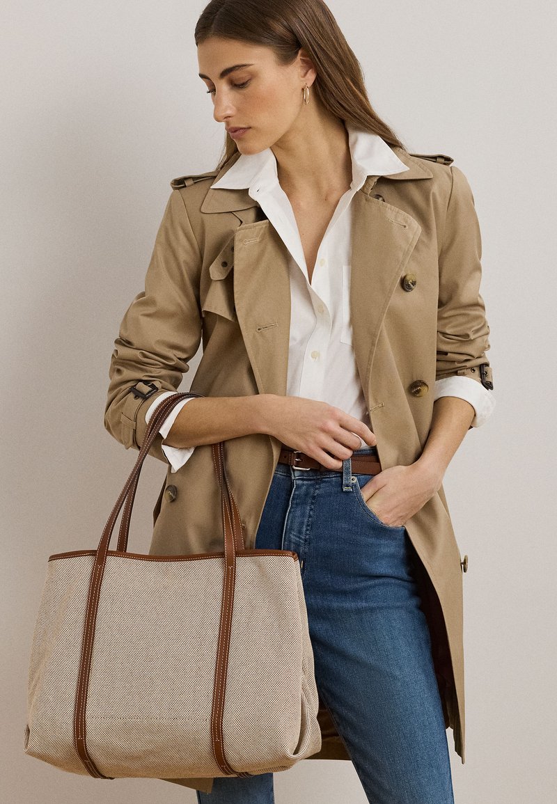 Vrouw draagt een beige trenchcoat en een wit overhemd, houdt een grote beige boodschappentas met bruine leren hengsels vast, staande tegen een effen achtergrond.
