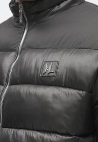 Jacheta neagră puffer are un guler înalt, design matlasat, textură lucioasă și un fermoar argintiu; include o mică etichetă cu logo pe piept.