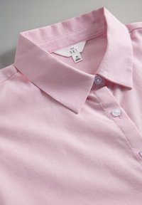 Svijetloružičasta košulja s ovratnikom i bijelim gumbima, vidljivom oznakom veličine "M" na neutralnoj pozadini.