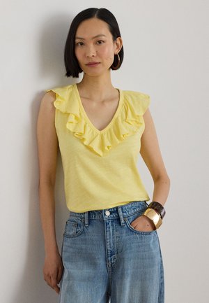 Γυναίκα που φοράει κίτρινο αμάνικο τοπ με βολάν, μπλε τζιν, μεγάλους κρίκους για σκουλαρίκια και φαρδιά βραχιόλια, με το ένα χέρι στην τσέπη της.