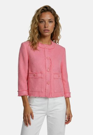 Femme aux cheveux blonds ondulés portant une veste rose texturée boutonnée avec deux poches devant et un pantalon blanc sur un fond uni.