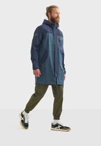 Un homme avec une barbe marche, portant une veste à capuche deux tons et un pantalon cargo, associés à des baskets sportives, sur un fond uni.