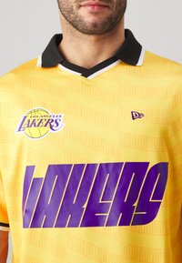 Gelbes Basketball-Trikot mit schwarzem Kragen, lila "LAKERS" Schriftzug und einem "LOS ANGELES LAKERS" Logo mit einer Basketballgrafik.