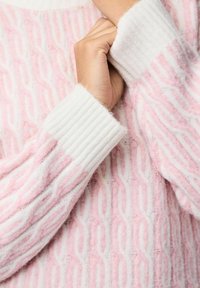 Rosa och vit texturerad tröja med flätmönster, med ribbade vita muddar och ett mjukt, lurvigt tyg.