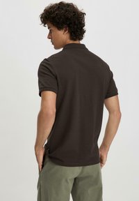 Brown polo shirt with a classic collar, short sleeves, and textured fabric. Worn with light green pants, viewed from the back.