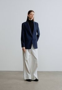 Granatowa marynarka na miarę z klapami z wycięciem, noszona na czarnym golfie, zestawiona z białymi spodniami o szerokich nogawkach i czarnymi butami.