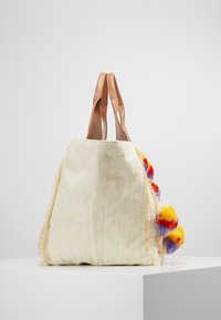 Torba typu tote w kremowym materiale z teksturowanym wzorem, z brązowymi skórzanymi uchwytami i kolorowymi pomponami w czerwonym, żółtym i niebieskim na jednej stronie.