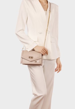 Donna in un completo bianco che tiene una borsa a tracolla rosa chiaro con catena dorata e chiusura a scatto.