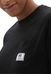 T-shirt en coton noir avec un col rond, doté d'une poche sur la poitrine gauche et d'un patch logo Vans blanc. Texture lisse, manches courtes.