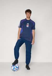 Conjunto desportivo azul-marinho com pormenores em vermelho e azul; a t-shirt de mangas curtas apresenta um logótipo; calças de fato de treino combinando, calçado preto e uma bola de futebol colorida.