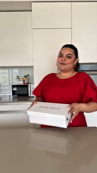 Fehér cipősdoboz, rajta fekete "ANNA FIELD" felirattal, egy vörös felsőt viselő személy által tartva, modern konyha háttérben.