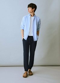 Camicia azzurra chiara a maniche lunghe sopra una t-shirt bianca, abbinata a pantaloni jogger scuri e scarpe slip-on marroni. Tessuto morbido, vestibilità comoda, design minimal.