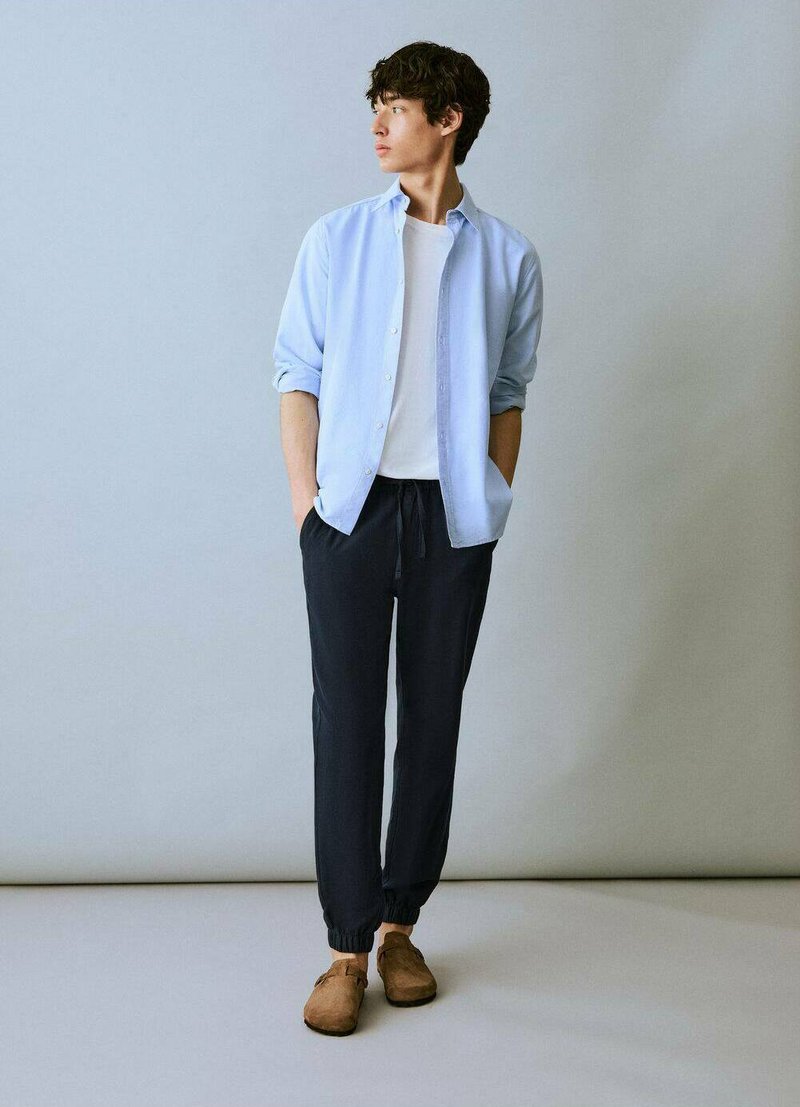 Camicia azzurra chiara a maniche lunghe sopra una t-shirt bianca, abbinata a pantaloni jogger scuri e scarpe slip-on marroni. Tessuto morbido, vestibilità comoda, design minimal.