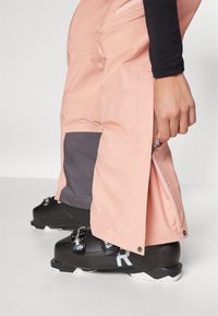 Light pink ski pants with gray accents, featuring side zippers and a sleek, waterproof fabric texture, paired with black ski boots.