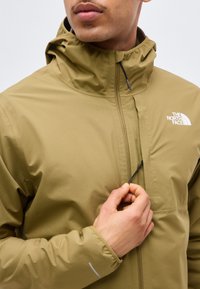 Άτομο που κλείνει με φερμουάρ το ελιάς πράσινο μπουφάν The North Face με ψηλό γιακά και ανακλαστική λεπτομέρεια στο μανίκι, σε ουδέτερο φόντο.