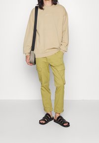 Sudadera beige de manga larga, pantalones cargo oliva con bolsillos laterales y sandalias negras con tiras. Diseño simple y funcional.