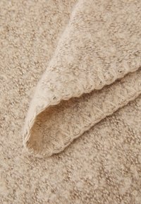 Tissu en tricot beige avec une surface douce et texturée. Un coin replié, révélant un bord festonné avec un motif de point décoratif.